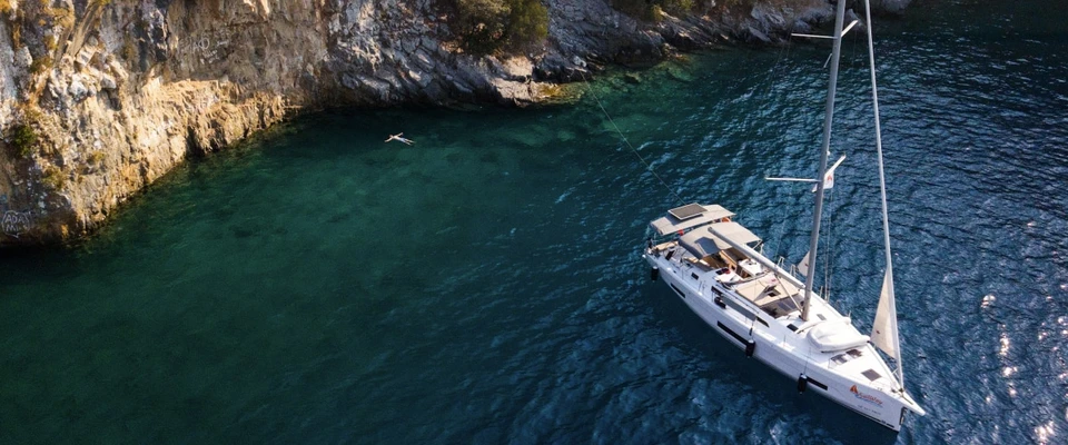 Aerial view of a sailboat at anchor in a turquoise bay beside a rocky coast.