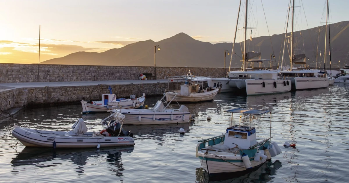 Boats moored in a Mediterranean harbour at sunset, with hills visible in the background.