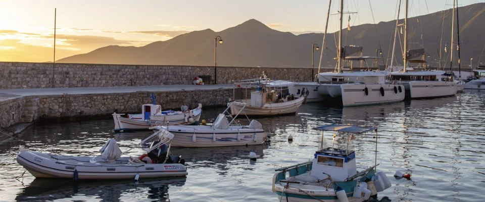 Boats moored in a Mediterranean harbour at sunset, with hills visible in the background.