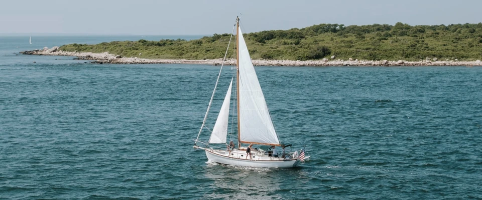 Sailboat cruising through clear turquoise coastal water near a green shoreline.