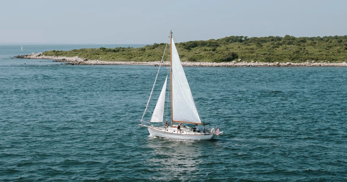 Sailboat cruising through clear turquoise coastal water near a green shoreline.