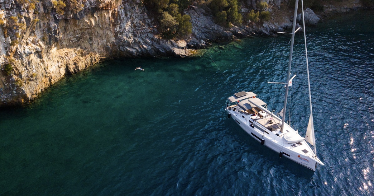 Aerial view of a sailboat at anchor in a turquoise bay beside a rocky coast.