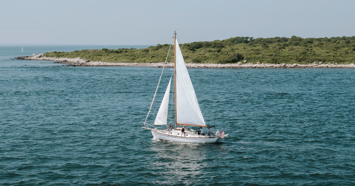 Sailboat cruising through clear turquoise coastal water near a green shoreline.